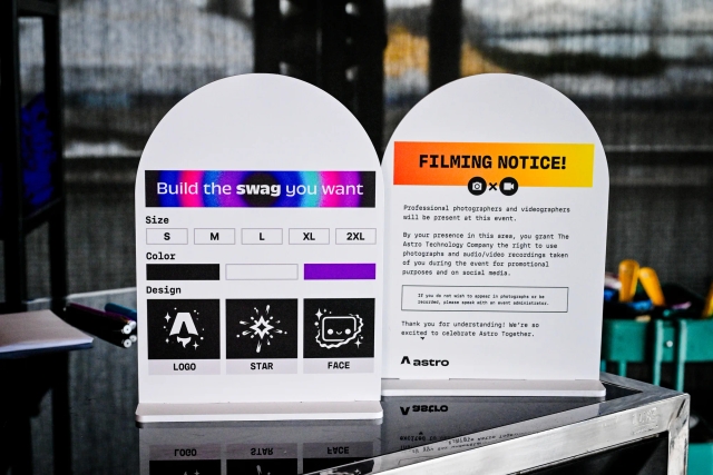 Two signs sitting atop a black bar. On the left, instructions to print your own t-shirt with sizes, colors, and designs. On the right, a filming notice in English.