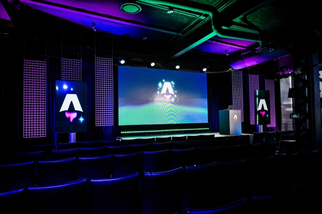 View from the back of the dark empty keynote presentation area. Rows empty black chairs. A vibrant large LED screen with the Astro Together logo on it. Two smaller portrait-oriented screens adorn the sides of the stage, also with the Astro Together logo.