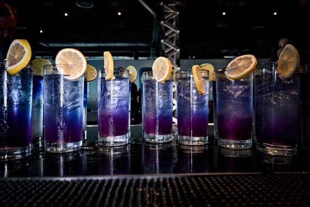 A dozen purple cocktails garnished with lime and lemon atop a black bar.