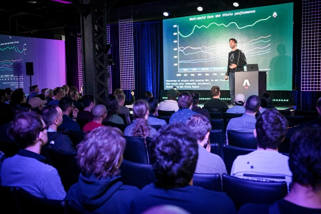 View from the back of the audience. Fred K. Schott is on stage with a slide depicting a graph, standing next to a branded Astro podium with a laptop on top of it.