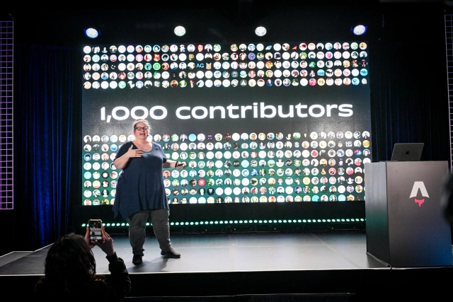 Sarah Rainsberger speaking from the left side of the stage, talking about how Docs has had 1,000 contributors to-date.