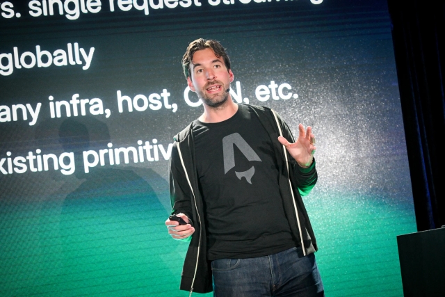 Fred K. Schott speaking on stage. He holds a clicker in his right hand and his left hand is open and slightly outstretched towards the crowd.